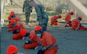 Detainees in orange jumpsuits sit in a holding area under the watchful eyes of Military Police at Camp X-Ray at Naval Base Guantanamo Bay, Cuba (Via Wikimedia Commons)